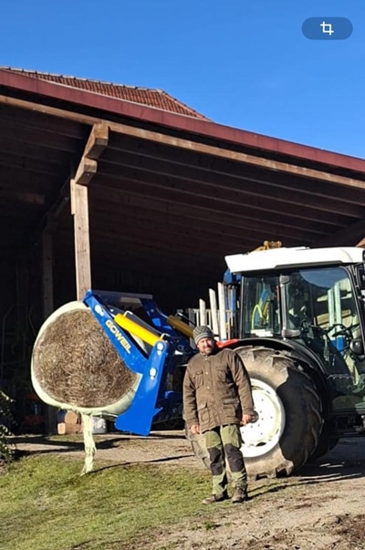 Traktor mit Frontlader hebt Heuballen vor Scheune, Landwirt arbeitet auf dem Bauernhof