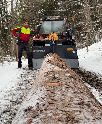 Forstarbeiter mit Traktor und Forstseilwinde zieht Baumstamm im verschneiten Wald