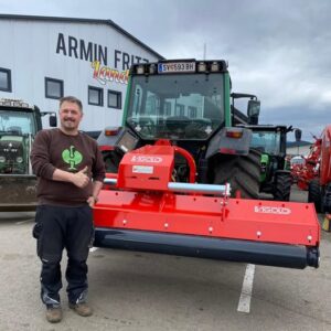 Mann gibt Daumen hoch neben einem Traktor mit rotem Vigolo-Anbaugerät vor dem Gebäude von Armin Fritz Landtechnik