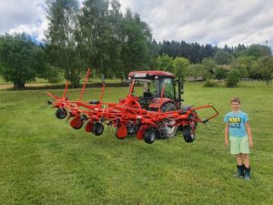 Roter Traktor mit Heuwender auf einer Wiese und ein daneben stehender Junge